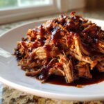 Close-up of tender, shredded Slow cooker pulled pork generously drizzled with dark BBQ sauce on a white plate.