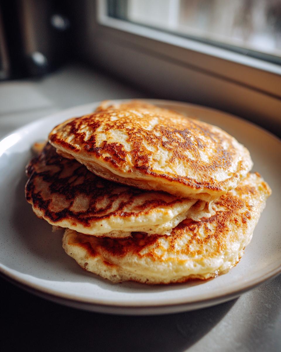 A stack of three fluffy sourdough pancakes with golden-brown, lacy edges served on a light plate near a window.
