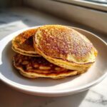 A stack of three perfectly golden brown sourdough pancakes sitting on a white plate, backlit by sunlight.