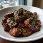 Close-up of tender, saucy crockpot steak bites piled high in a white bowl and topped with fresh chopped parsley.