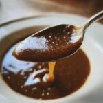 Close-up of thick, rich homemade brown gravy dripping from a silver spoon back into a white bowl.