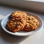 Three golden brown, textured oatmeal cookie treats resting on a small white plate, highlighted by natural sunlight.