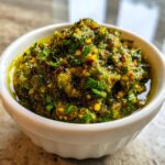 Close-up of thick, chunky green tomato salsa, vibrant green with visible herbs and chili flakes, served in a small white bowl.