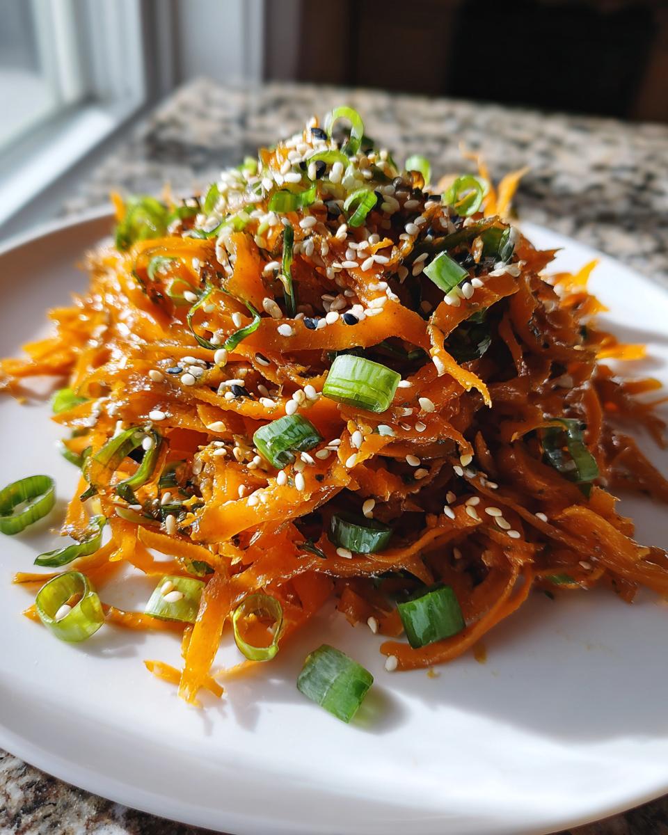 A mound of bright orange raw carrot salad, thinly shredded and coated in dressing, topped with sesame seeds and sliced green onions.