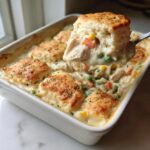 A spoonful of creamy chicken pot pie casserole filling with vegetables being lifted from a white baking dish topped with golden biscuits.