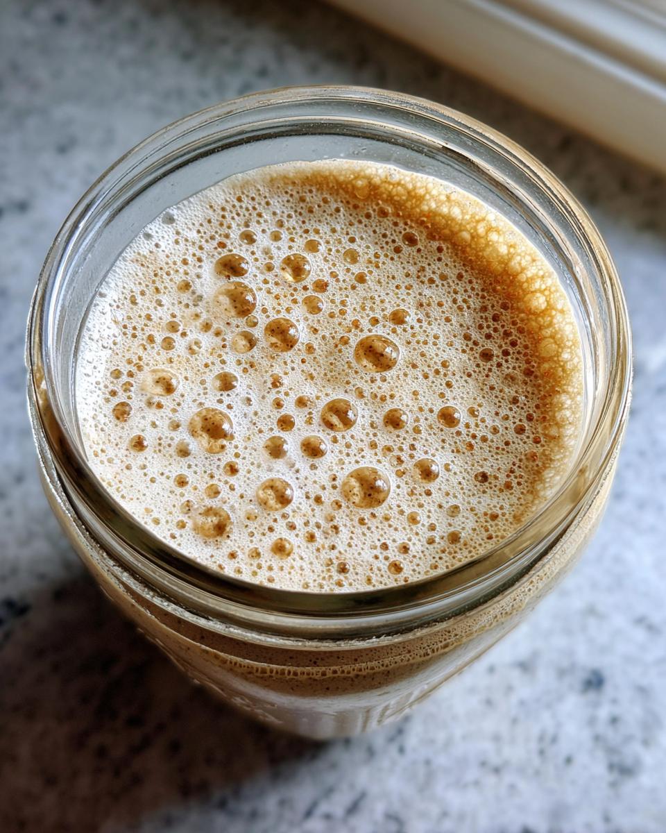 Overhead view of an active sourdough starter showing a thick layer of foam and bubbles in a glass jar.