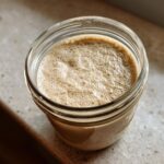Close-up of a bubbly, active sourdough starter in a clear glass jar, showing signs of successful fermentation.