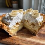 A cross-section view of a rich banana cream pie showing layers of crust, custard, bananas, and whipped cream topping.