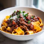 A close-up of tender shredded beef ragu served over wide pappardelle noodles, topped with Parmesan and parsley.