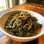 A close-up of tender, dark green collard greens served in a white bowl with savory broth and shredded smoked meat.