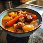 A close-up of a hearty bowl of pork stew featuring tender meat chunks, potatoes, and bright orange carrots in a rich broth.
