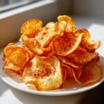 A mound of golden, crispy homemade potato chips piled high on a white plate, catching bright sunlight.