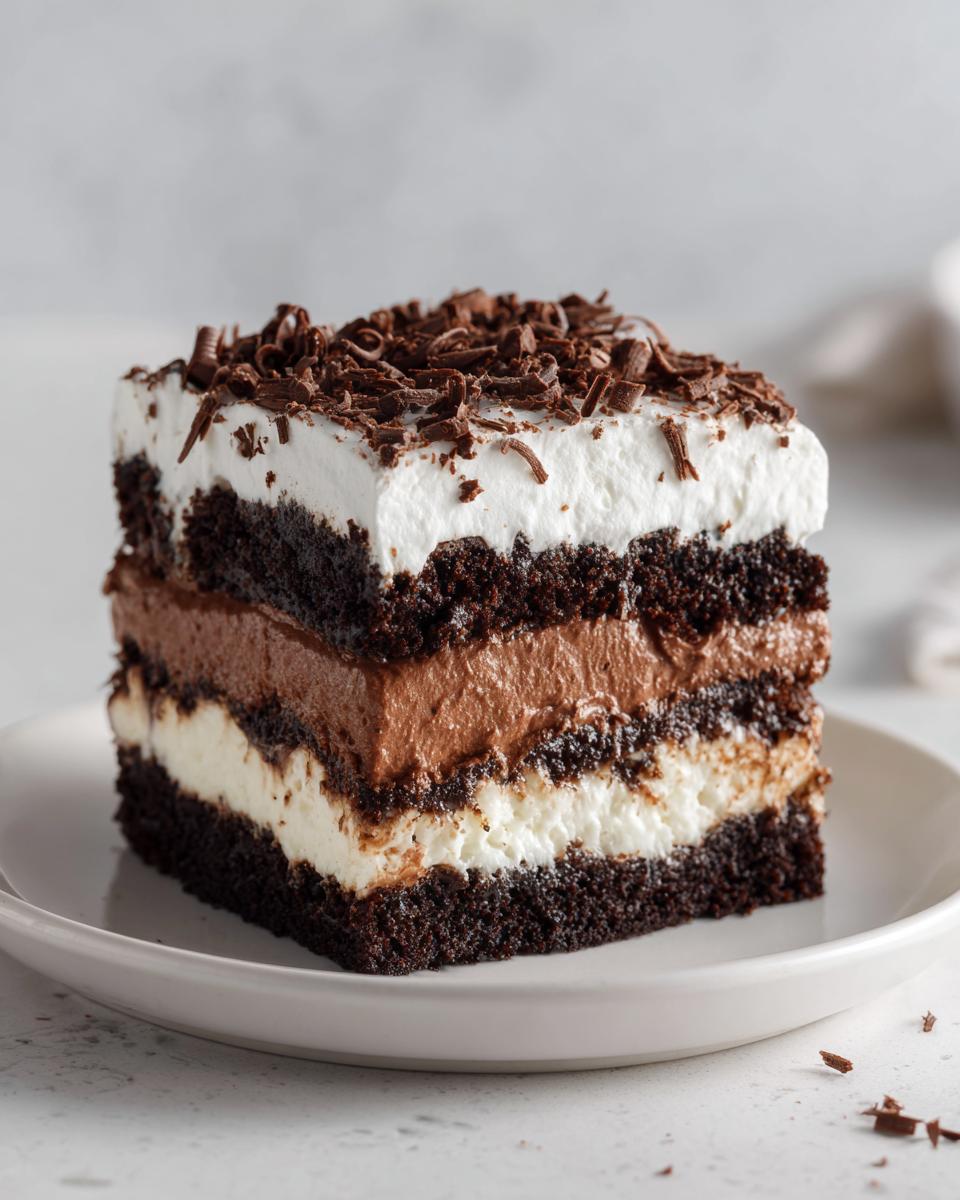 A decadent square slice of brownie refrigerator cake with layers of dark cake, chocolate mousse, and white cream, topped with chocolate shavings.