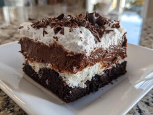 A rich slice of brownie refrigerator cake showing layers of dark brownie, white cream, chocolate pudding, and whipped topping with chocolate shavings.