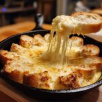 A slice of toasted bread is lifted from a skillet of hot, bubbly cheese dip, showing a dramatic cheese pull.