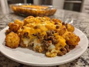 A close-up serving of cheesy tater tot casserole featuring crispy tater tots, ground beef, and melted cheddar cheese on a white plate.