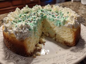 A slice removed from a festive Christmas tree cheesecake topped with green frosting, sprinkles, and piped whipped cream.