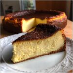 A close-up shot of a creamy slice of Basque burnt cheesecake on a white plate, with the rest of the cake blurred in the background.