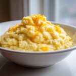 A close-up of a white bowl filled with creamy, yellow cheese grits topped with shredded cheddar cheese.