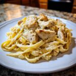 A close-up shot of creamy chicken alfredo pasta with chunks of seasoned chicken served on a white plate.