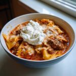 A close-up of a bowl of creamy lasagna soup featuring pasta, meat sauce, and a large dollop of ricotta cheese.