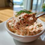 A spoonful of creamy smoked salmon dip, garnished with fresh dill, being lifted from a white bowl.