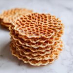 A close-up stack of golden brown, crisp pizzelle cookies showing their honeycomb pattern, resting on a white marble surface.