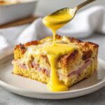 A square slice of eggs benedict casserole topped with ham and cheese, being drizzled with bright yellow hollandaise sauce from a spoon.