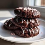 A stack of three fudgy double chocolate peppermint cookies drizzled with white icing and topped with crushed candy canes.