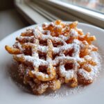 A golden brown, freshly fried funnel cake recipe creation generously dusted with white powdered sugar on a white plate.
