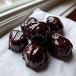 A small pile of glossy, dark chocolate covered cherries resting on a white cloth near a window.