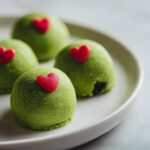 Four bright green Grinch Oreo balls topped with small red candy hearts, resting on a white plate.