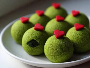 Several bright green, textured Grinch Oreo Balls topped with small red hearts, arranged on a white plate.