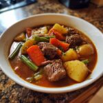 Close-up of a white bowl filled with rich vegetable beef soup, featuring large chunks of beef, potatoes, carrots, and green beans.