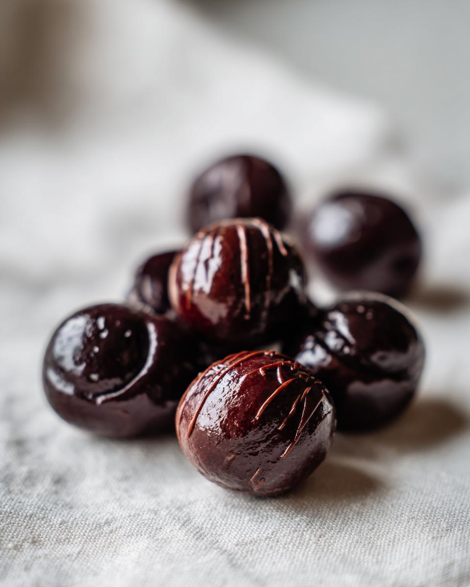 A small pile of glossy, dark chocolate covered cherries, one featuring a drizzle decoration, resting on a light linen surface.