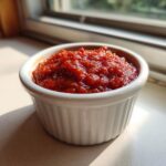 A small white ramekin filled with thick, vibrant red homemade cocktail sauce, sitting on a counter near a window.