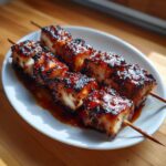 Two skewers of juicy, perfectly cooked chicken skewers coated in a dark, sticky glaze resting on a white plate.