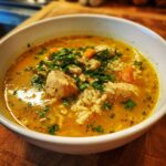 Close-up of a white bowl filled with vibrant yellow Lemon Ginger Turmeric Chicken and Rice Soup, topped with chicken chunks, rice, carrots, and parsley.