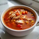 Close-up of a steaming white bowl filled with rich, red Maryland crab soup, topped with large flakes of white crab meat, carrots, and corn.