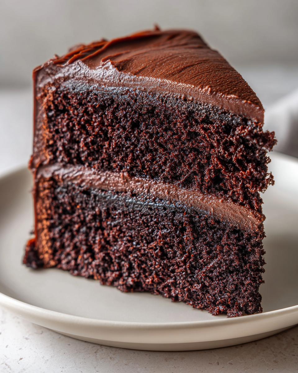 Close-up of a tall, dark slice of moist chocolate cake layered with thick chocolate frosting.