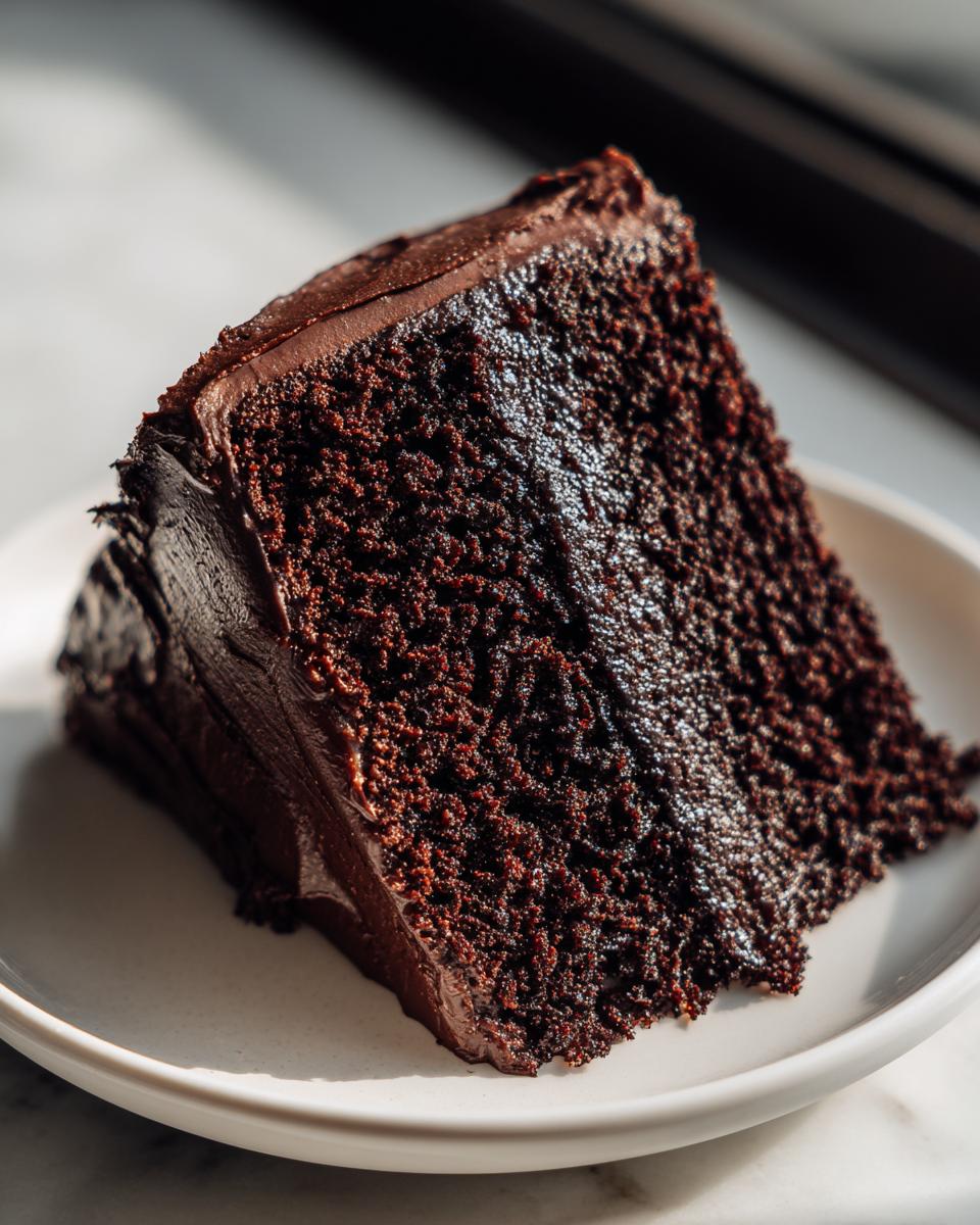 Close-up of a rich, dark slice of moist chocolate cake covered in thick chocolate frosting.
