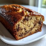 A close-up of a freshly baked, moist banana bread loaf studded with chocolate chips, resting on a white plate.