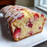Close-up of a moist strawberry bread loaf drizzled with white vanilla icing on a white plate.