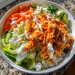 A vibrant bowl featuring shredded buffalo chicken, rice, lettuce, carrots, blue cheese, and a ranch dressing drizzle.