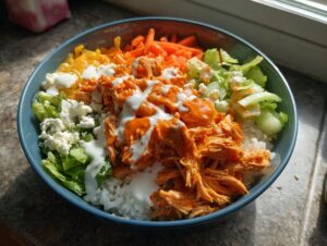 A vibrant bowl featuring shredded buffalo chicken over rice, topped with ranch dressing, lettuce, carrots, and blue cheese crumbles.