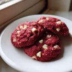 A close-up of three vibrant red velvet cake mix cookies studded with white chocolate chips on a white plate.