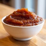 A close-up of thick, rich brown pear butter mounded high in a small white bowl on a wooden surface.