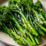 A close-up shot of bright green, roasted broccolini spears sprinkled with grated Parmesan cheese on a light plate.