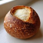 A close-up of a freshly baked, round bread bowl with a dark, crusty exterior and the soft white interior visible.