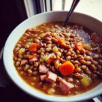 Close-up of a white bowl filled with savory 15 bean soup, featuring beans, diced carrots, celery, and chunks of ham.